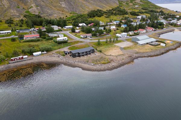 Aerial view of a coastal village with houses on a green hillside, a dark modern building on a rocky bay, and mountains in the background.