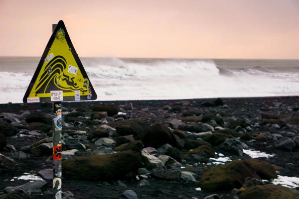 Una señal de advertencia amarilla con una ola estilizada arrastrando a una persona se encuentra en una playa rocosa negra con grandes olas rompiendo al fondo.
