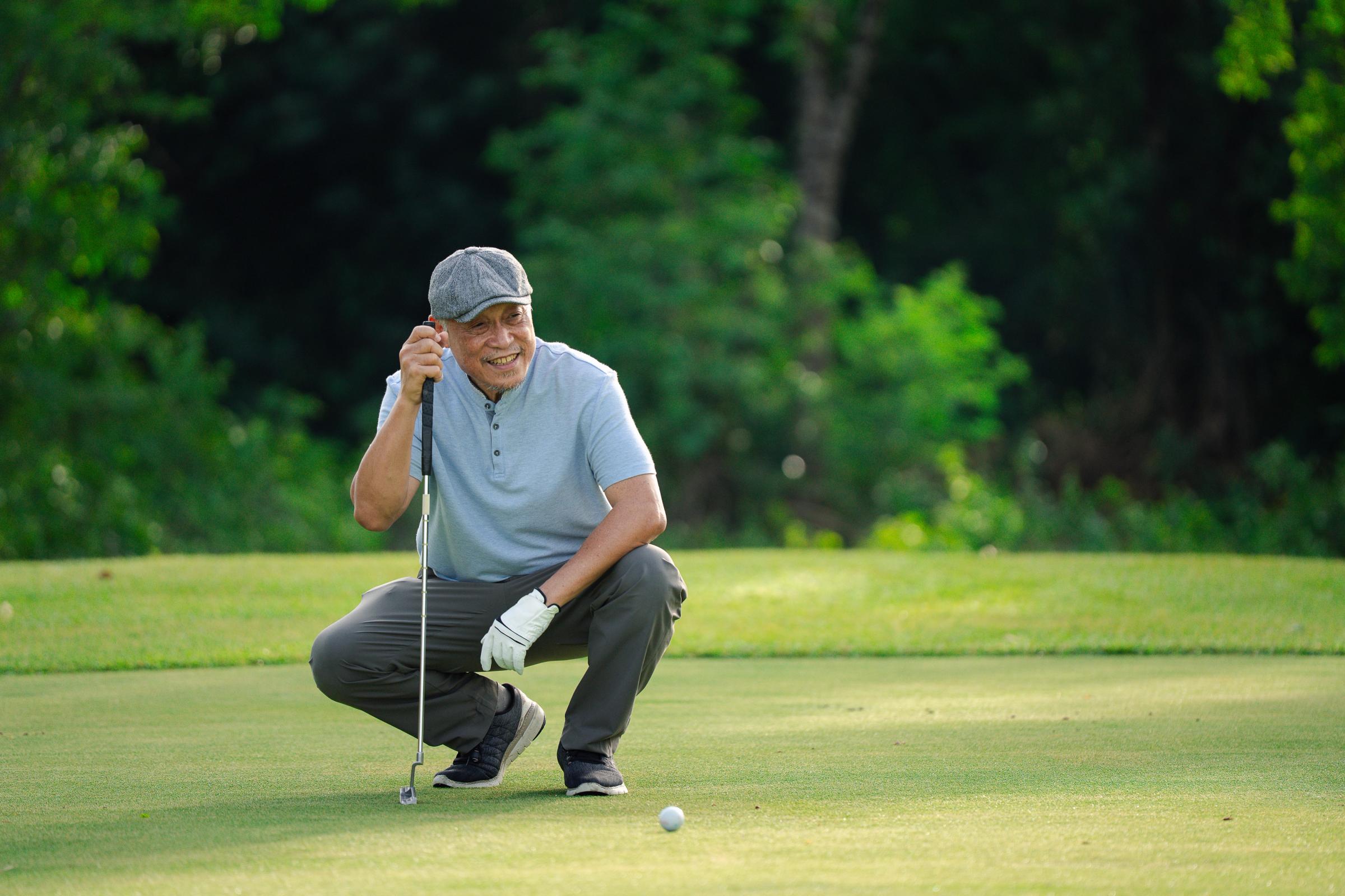 A smiling man in a flat cap crouches on a golf course, holding a putter near a golf ball.