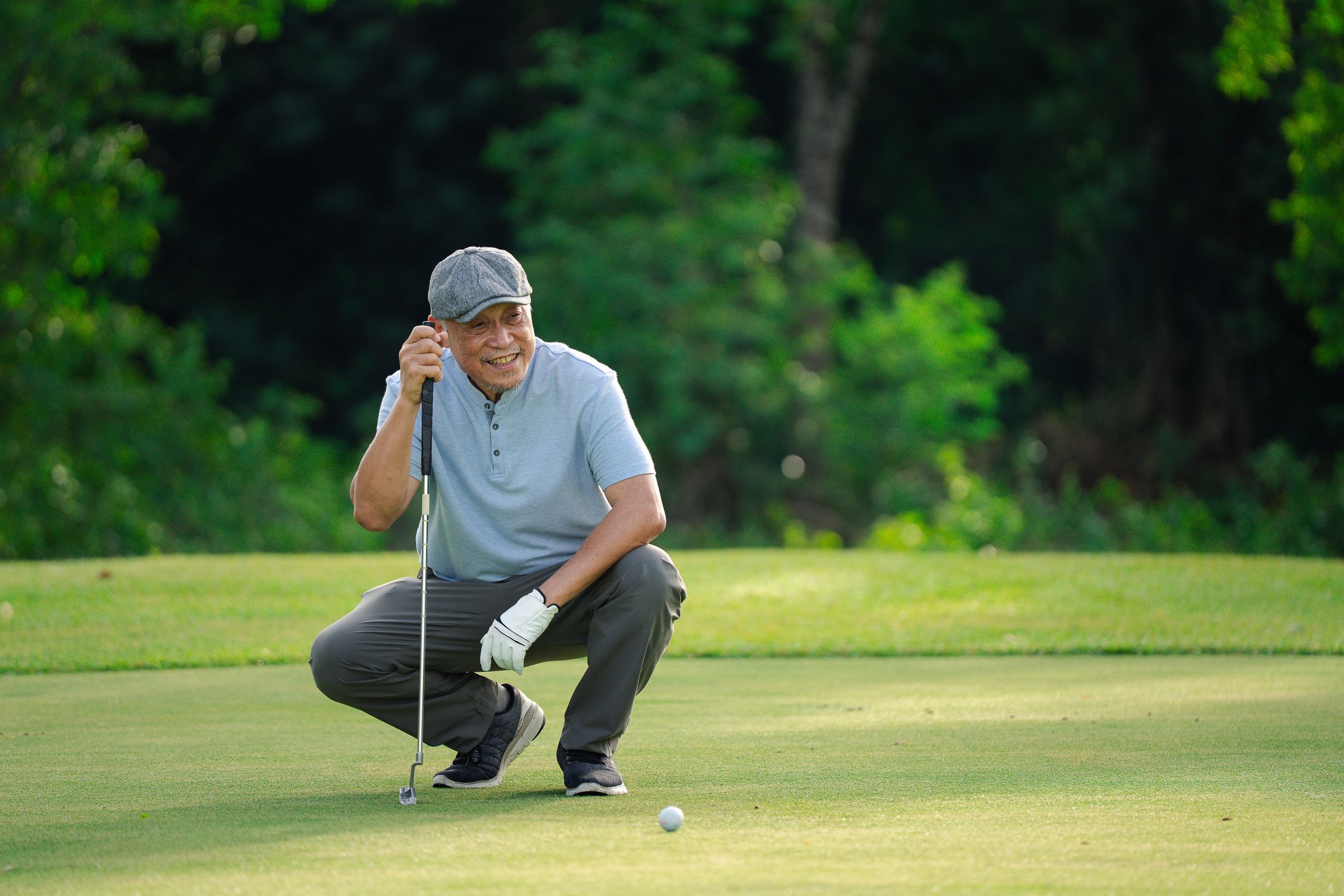 A smiling man in a flat cap crouches on a golf course, holding a putter near a golf ball.