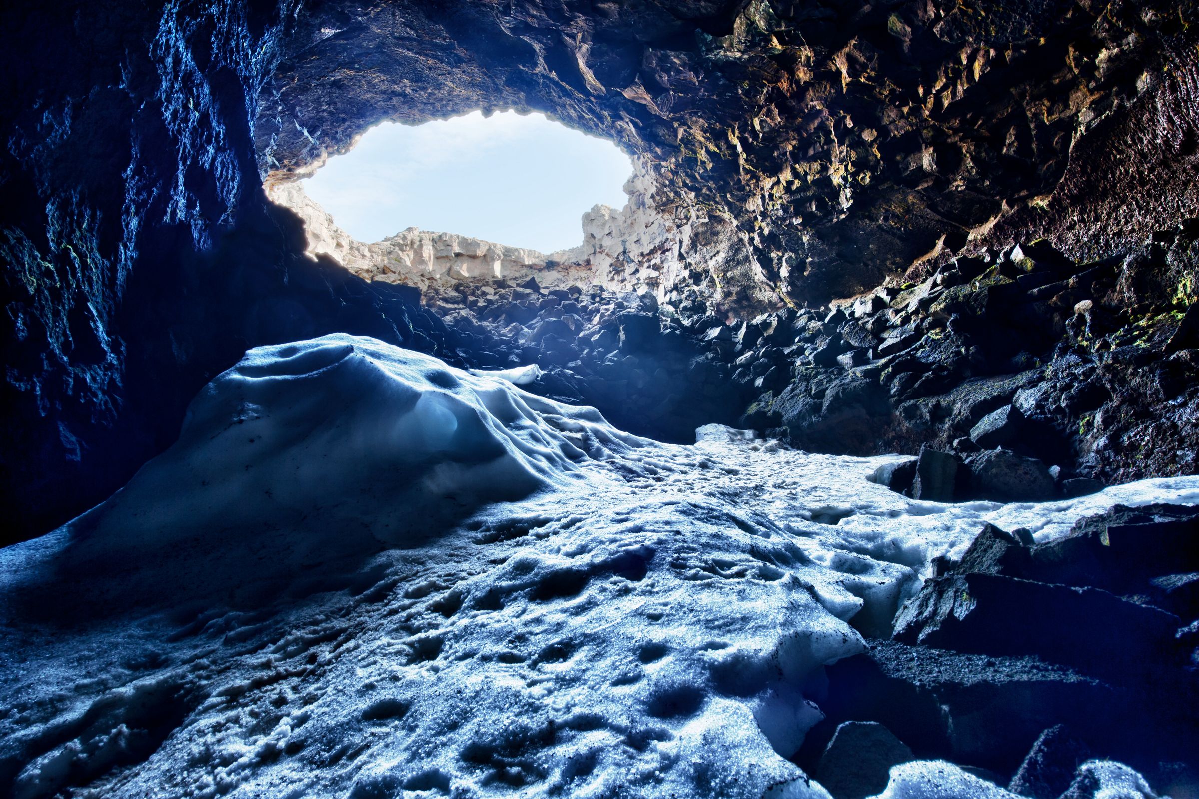Interior oscuro y rocoso de una cueva con un suelo de nieve y hielo tintados de azul, que conduce a una luminosa apertura hacia el cielo.