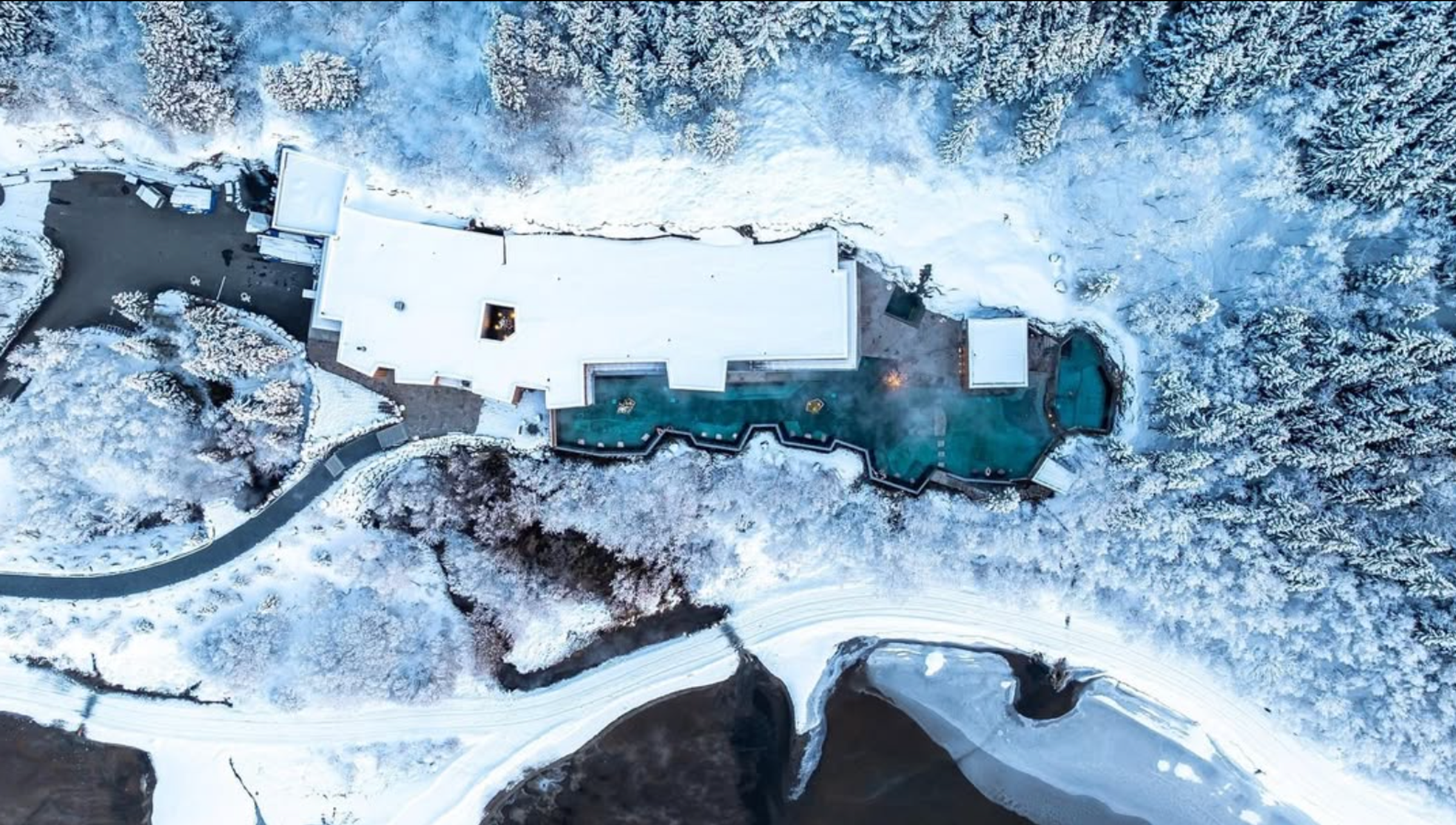 Aerial view of a snow-covered resort with steaming hot springs, surrounded by a winter forest and a river.