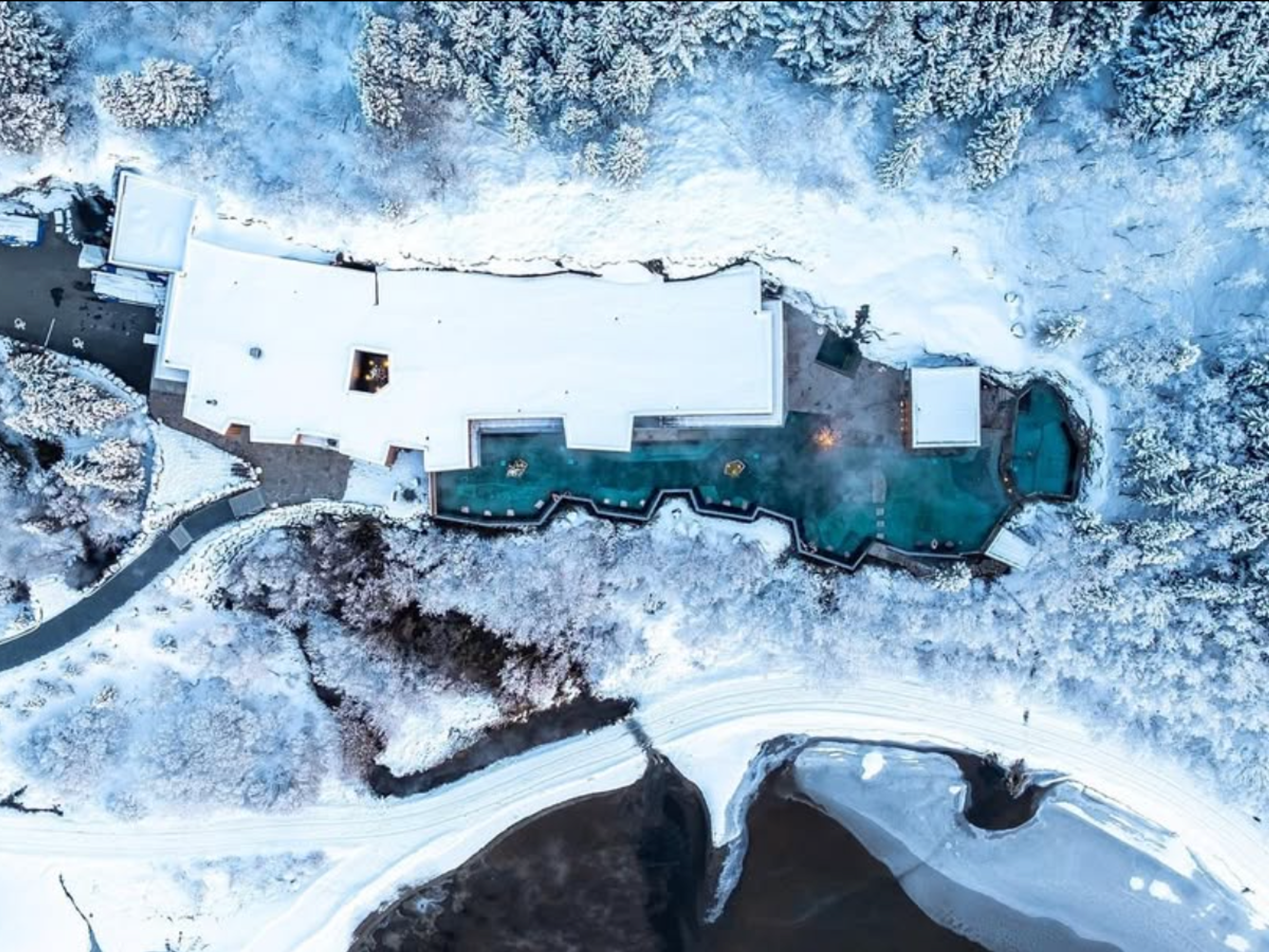 Aerial view of a snow-covered resort with steaming hot springs, surrounded by a winter forest and a river.
