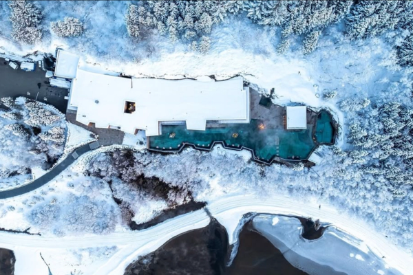 Vista aérea de un complejo turístico cubierto de nieve con aguas termales azules humeantes rodeado de un bosque invernal y un río parcialmente congelado.
