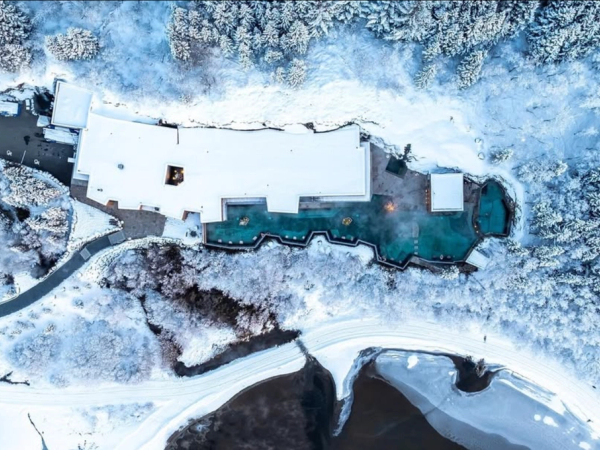 Vista aérea de un complejo turístico cubierto de nieve con aguas termales humeantes, rodeado de un bosque invernal y un río.