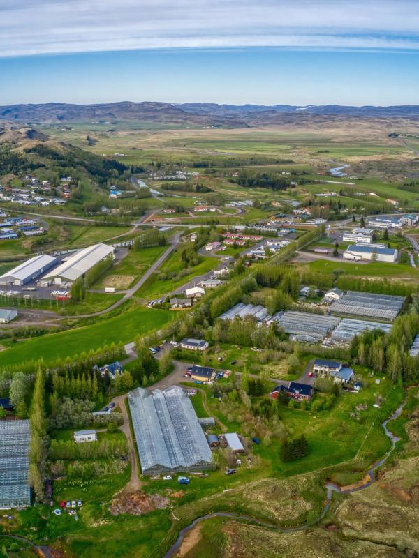 Aerial view of a town in a green valley with many greenhouses, a winding river, and distant snow-capped mountains.
