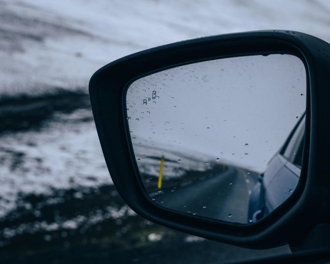 Espejo retrovisor lateral de un Mitsubishi Eclipse Cross que refleja una carretera asfaltada, con gotas de lluvia en el cristal