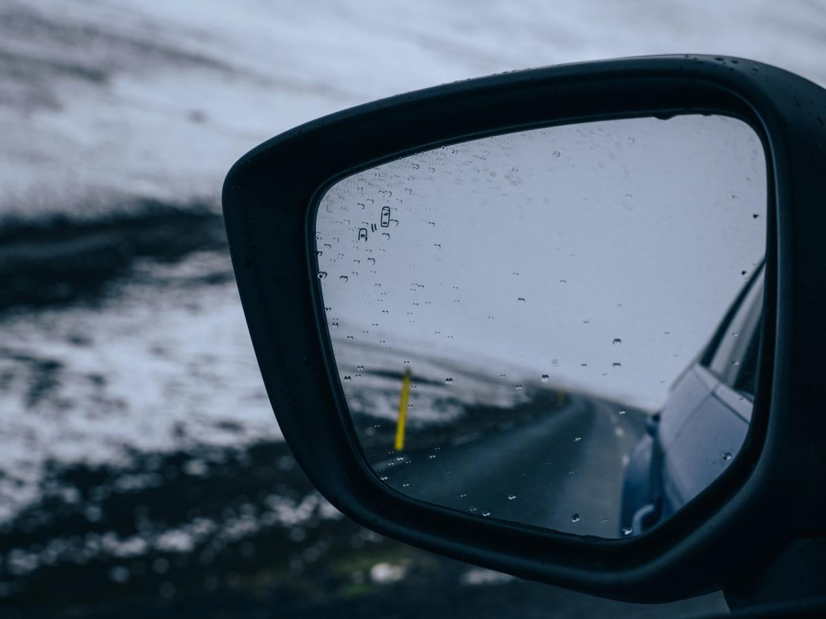 Vista del retrovisor del Mitsubishi Eclipse Cross Espejo retrovisor lateral de un Mitsubishi Eclipse Cross que refleja una carretera asfaltada, con gotas de lluvia en el cristal