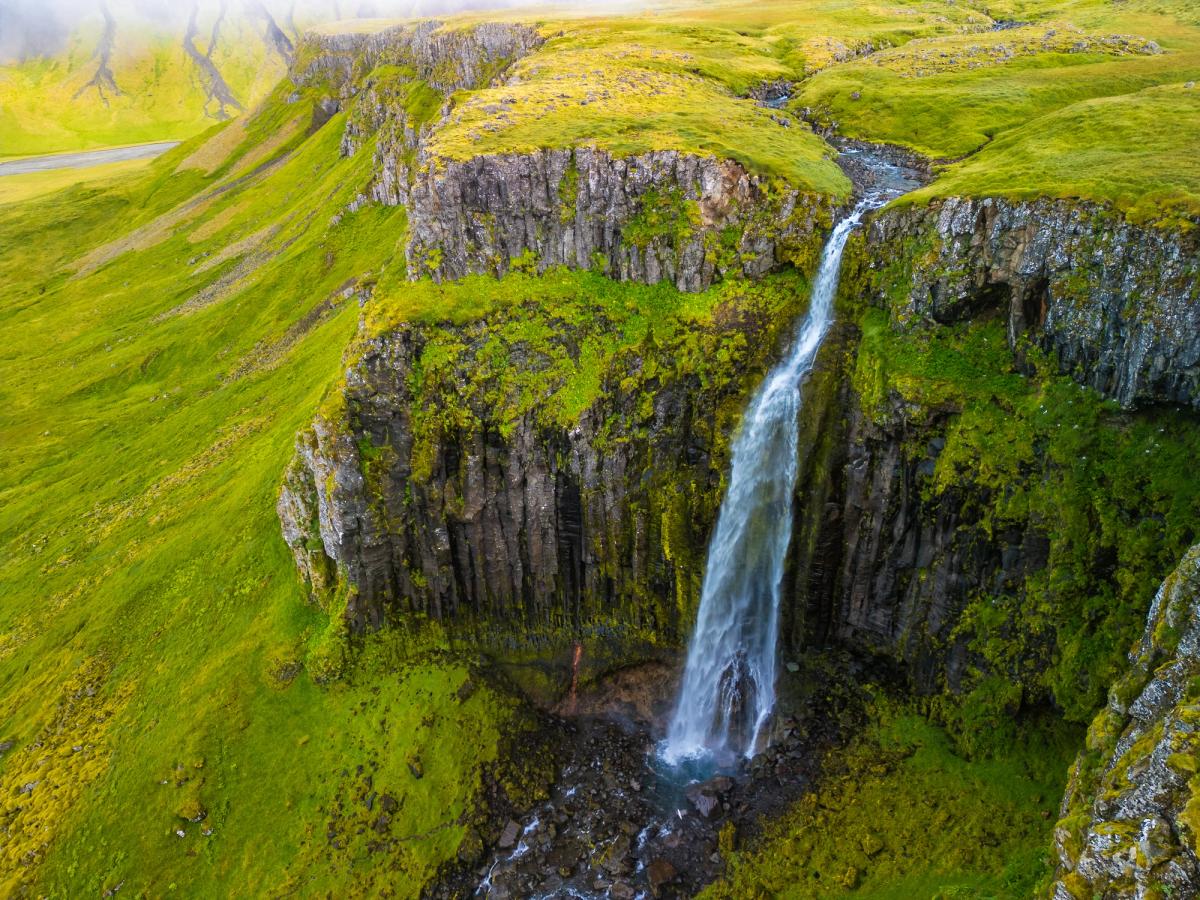 A tall waterfall cascades over mossy, dark cliffs amidst vibrant green hills.