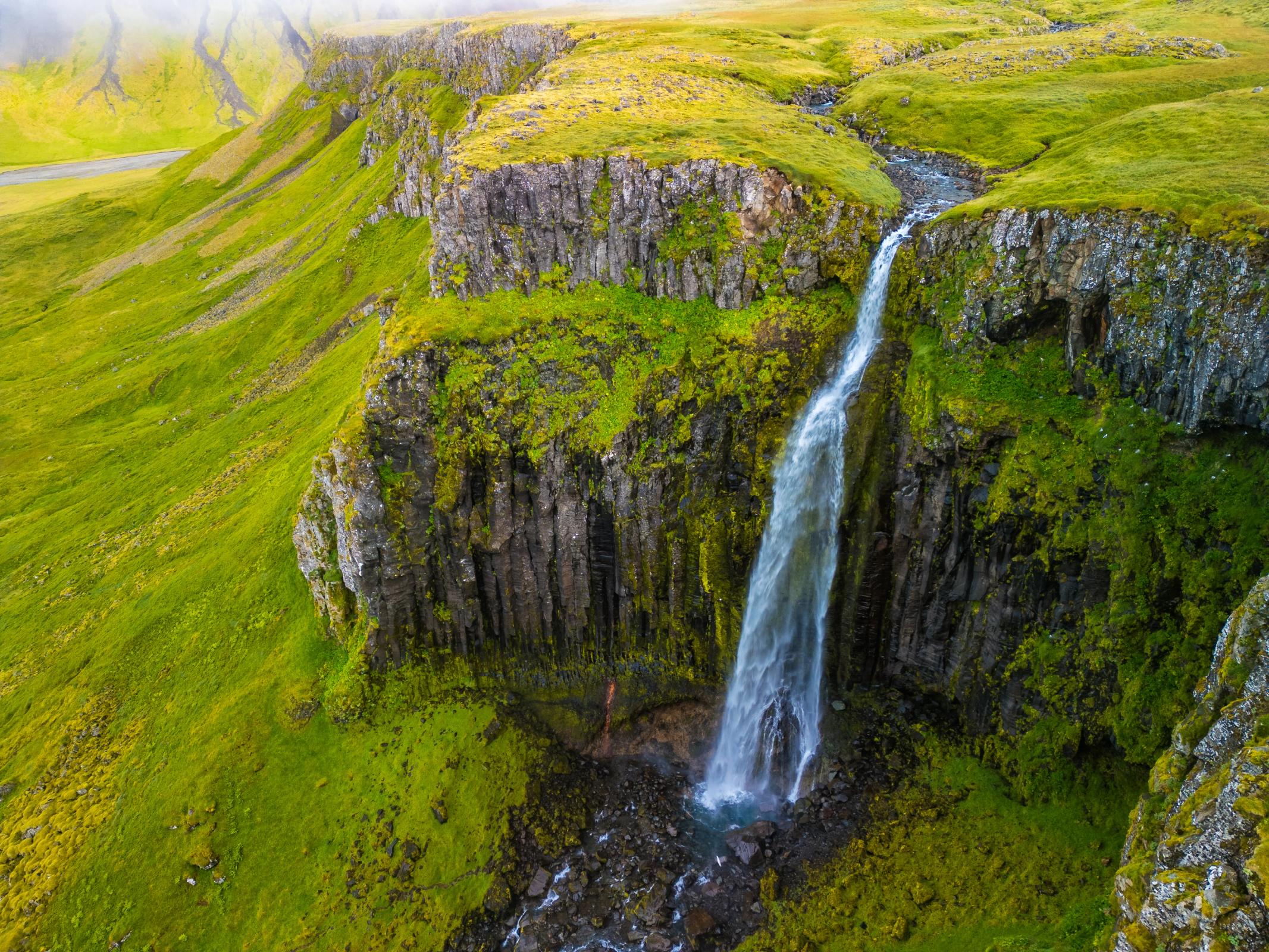 A tall waterfall plunges over a mossy columnar basalt cliff surrounded by bright green hills.