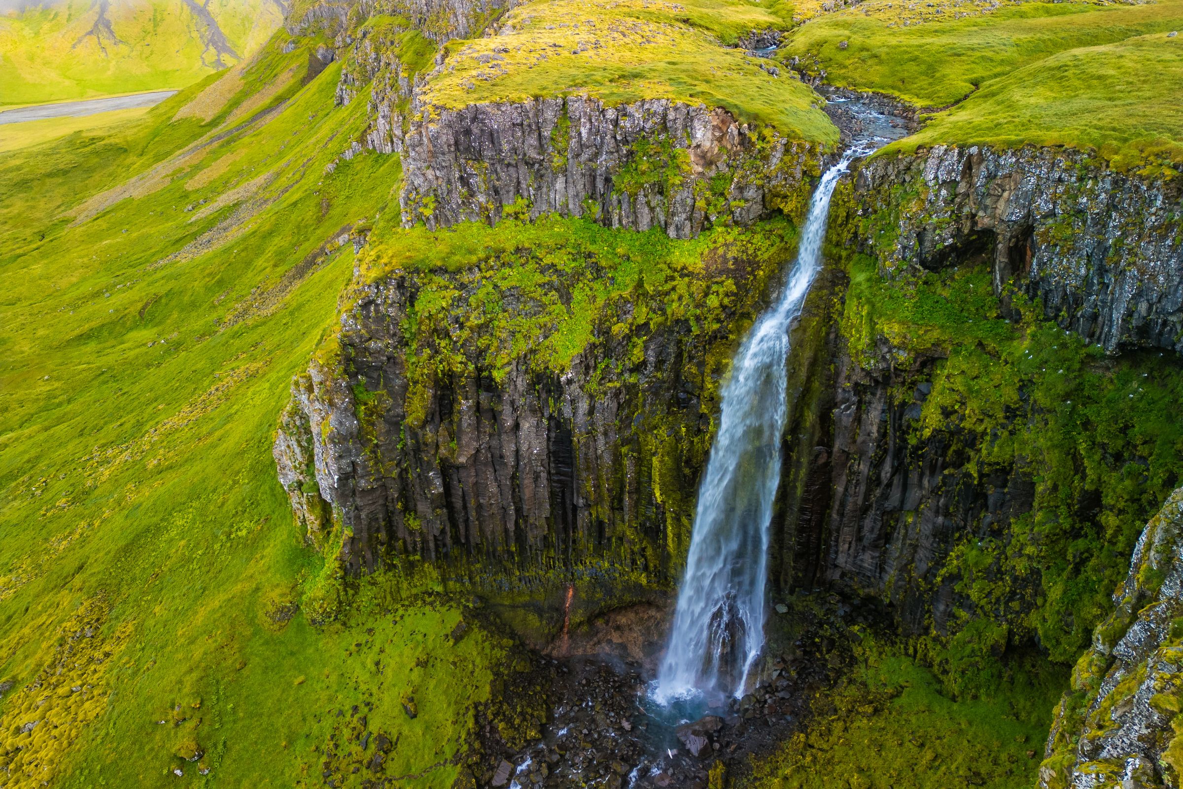 A tall waterfall plunges over a mossy columnar basalt cliff surrounded by bright green hills.