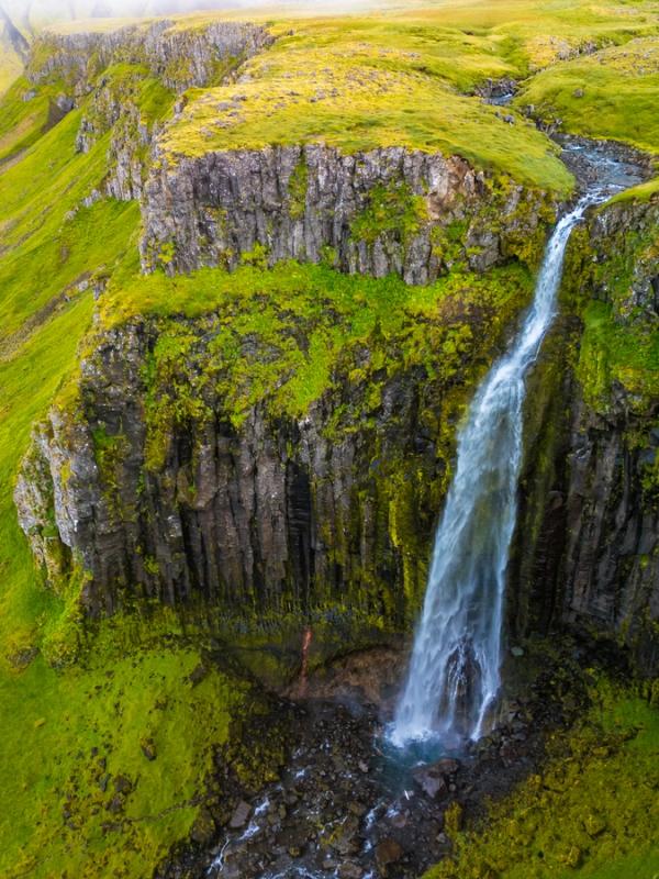 A tall waterfall cascades over mossy, dark cliffs amidst vibrant green hills.