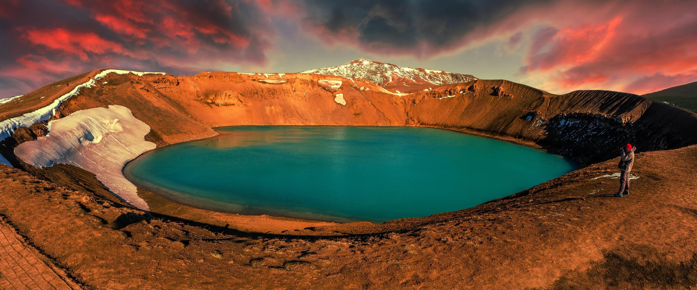 A person views a vibrant turquoise crater lake in a reddish volcanic landscape under a dramatic red sky.