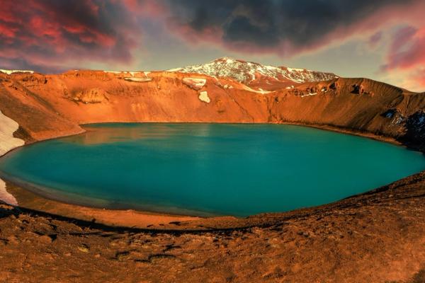 A person views a vibrant turquoise crater lake in a reddish volcanic landscape under a dramatic red sky.
