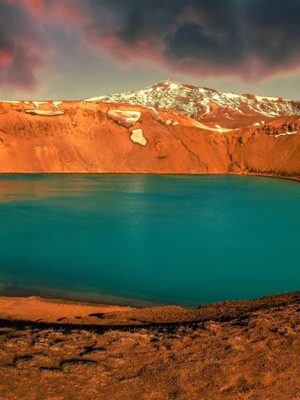 A person views a bright turquoise crater lake with snow-dusted reddish walls under a dramatic red sky.