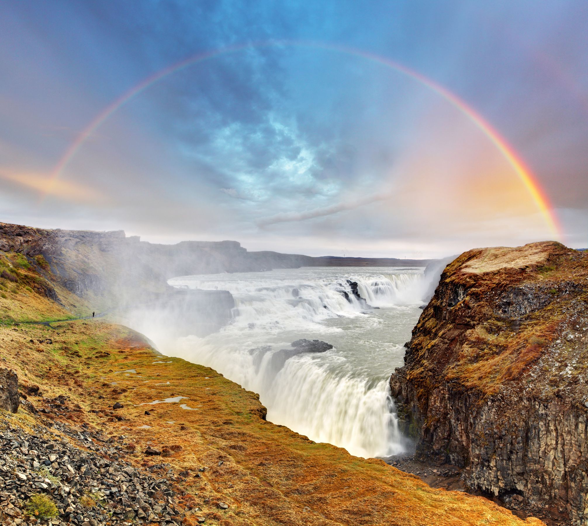 A wide rainbow arcs over a powerful waterfall cascading through a rocky canyon.