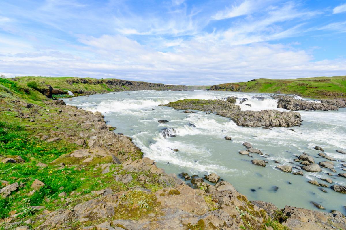 A wide river with multiple rapids and small waterfalls flowing through a rocky, grassy landscape under a blue sky.