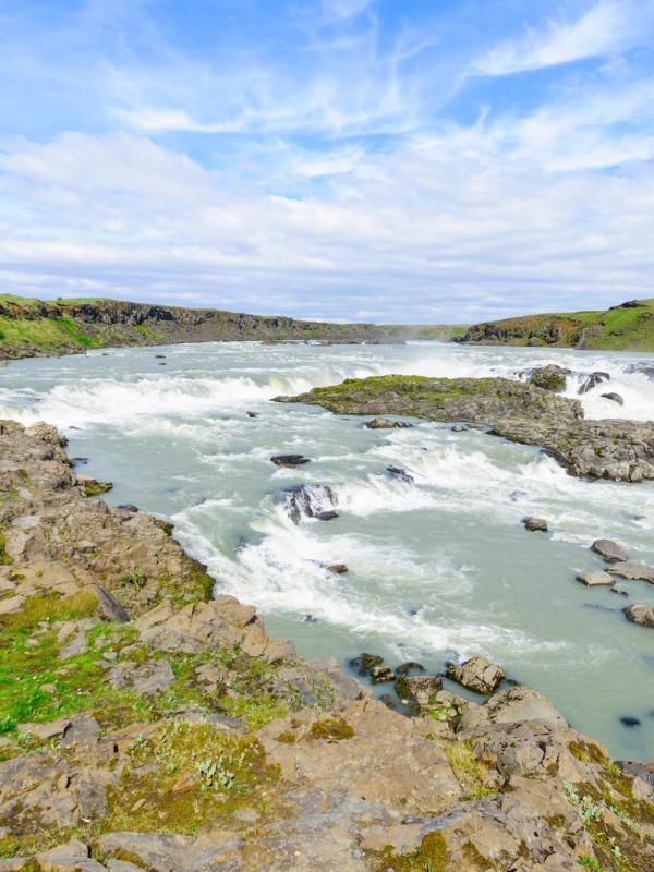 A wide river with multiple rapids and small waterfalls flowing through a rocky, grassy landscape under a blue sky.