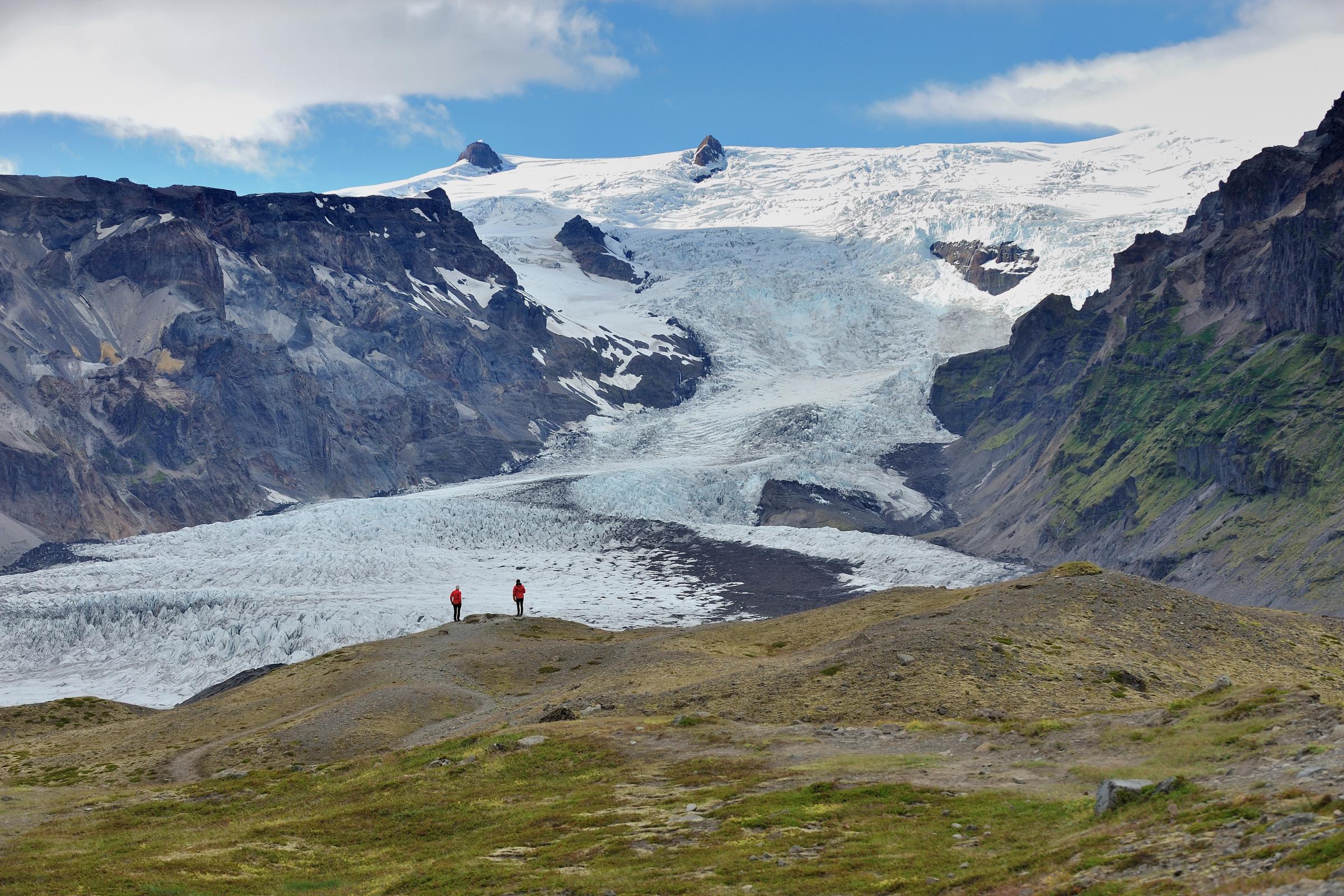 two people are standing on top of a hill in front of a large glacier .