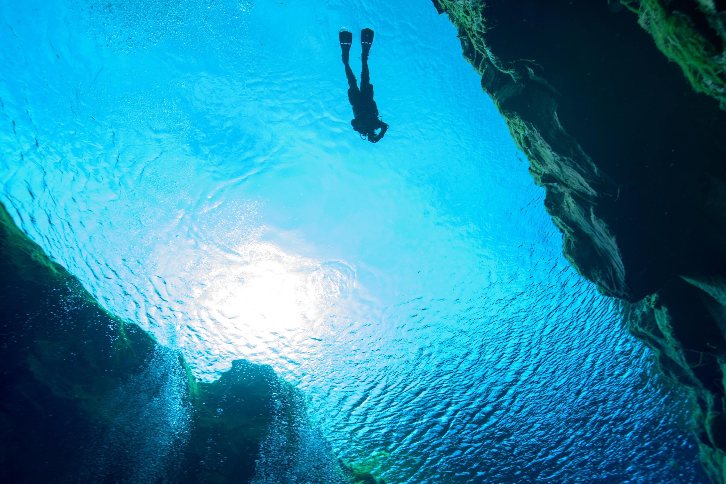 Vista submarina de un buzo silueteado contra la brillante superficie del agua, enmarcado por las oscuras paredes de una cueva con burbujas ascendiendo.