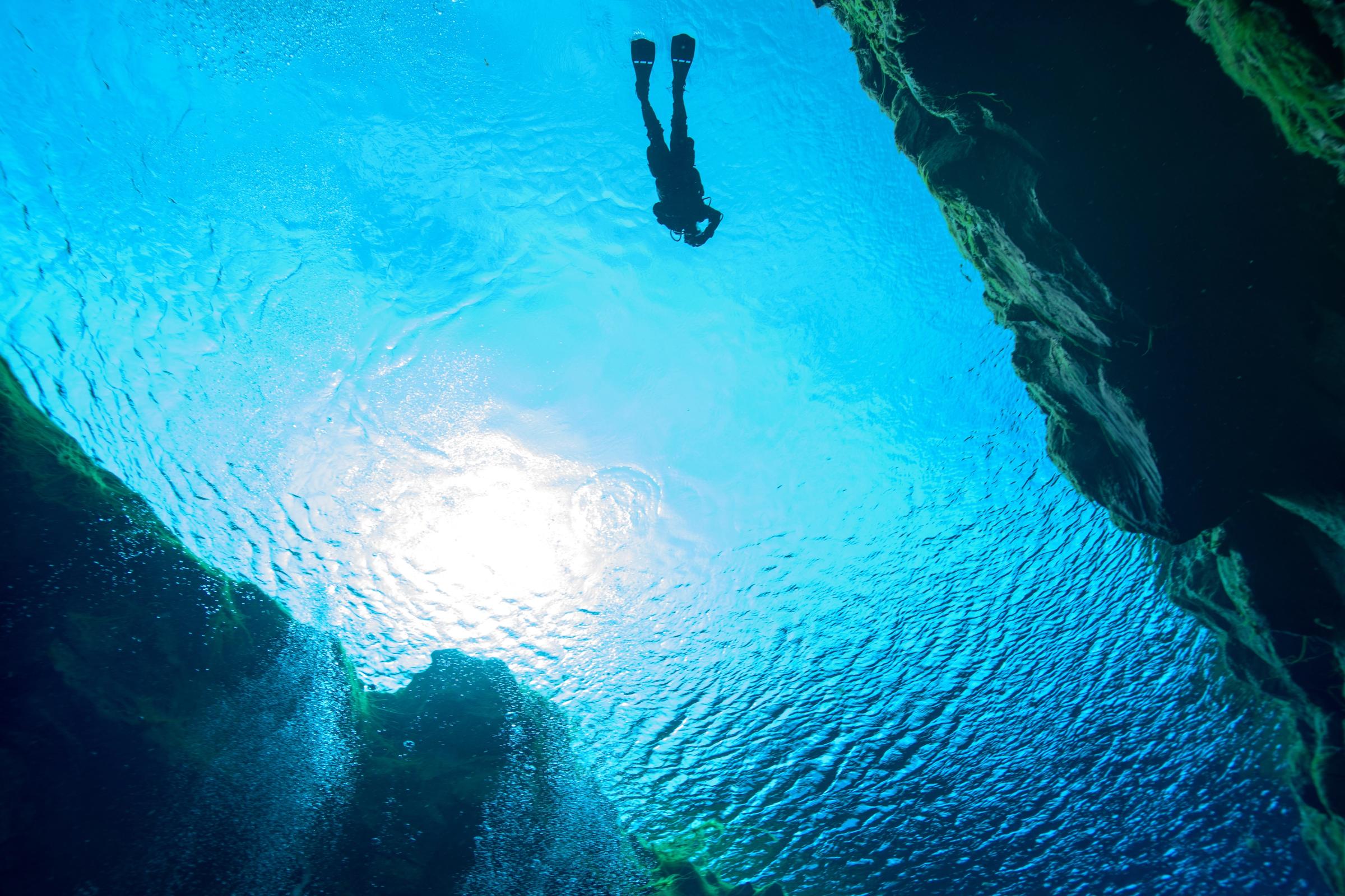Un buzo recortado contra la brillante superficie del agua en una cueva submarina, con burbujas que ascienden.