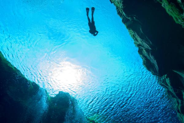 Underwater view of a diver silhouetted against the bright water surface, framed by dark cave walls with rising bubbles.