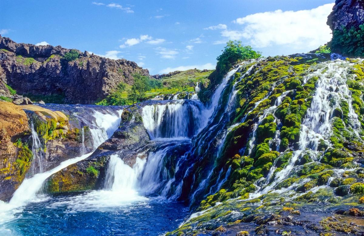 A waterfall cascades over moss-covered rocks into a blue pool, under a bright blue sky.