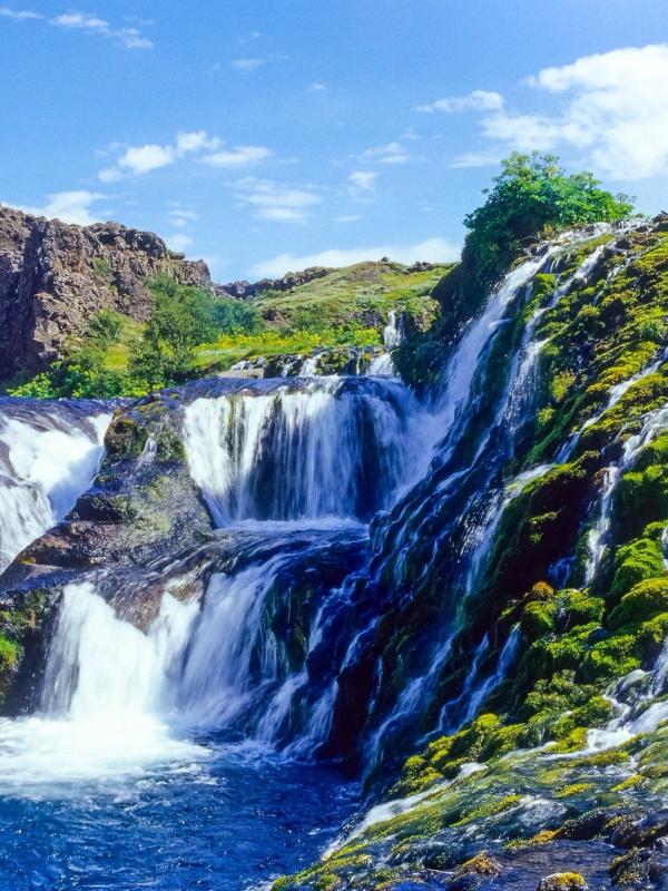 A waterfall cascades over moss-covered rocks into a blue pool, under a bright blue sky.