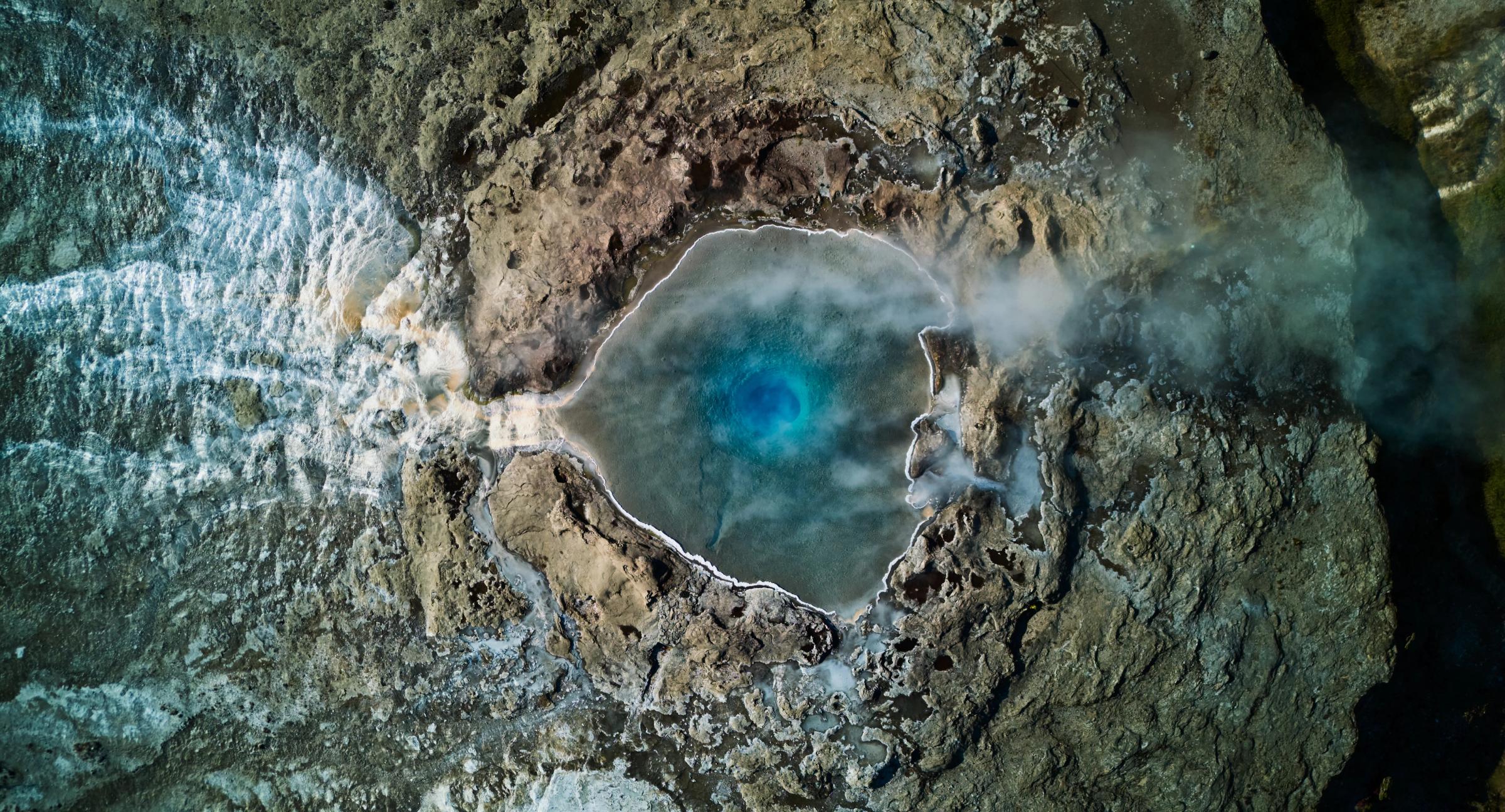 Vista aérea de una vibrante poza termal azul que humea entre un terreno rocoso, con una potente cascada a su izquierda.