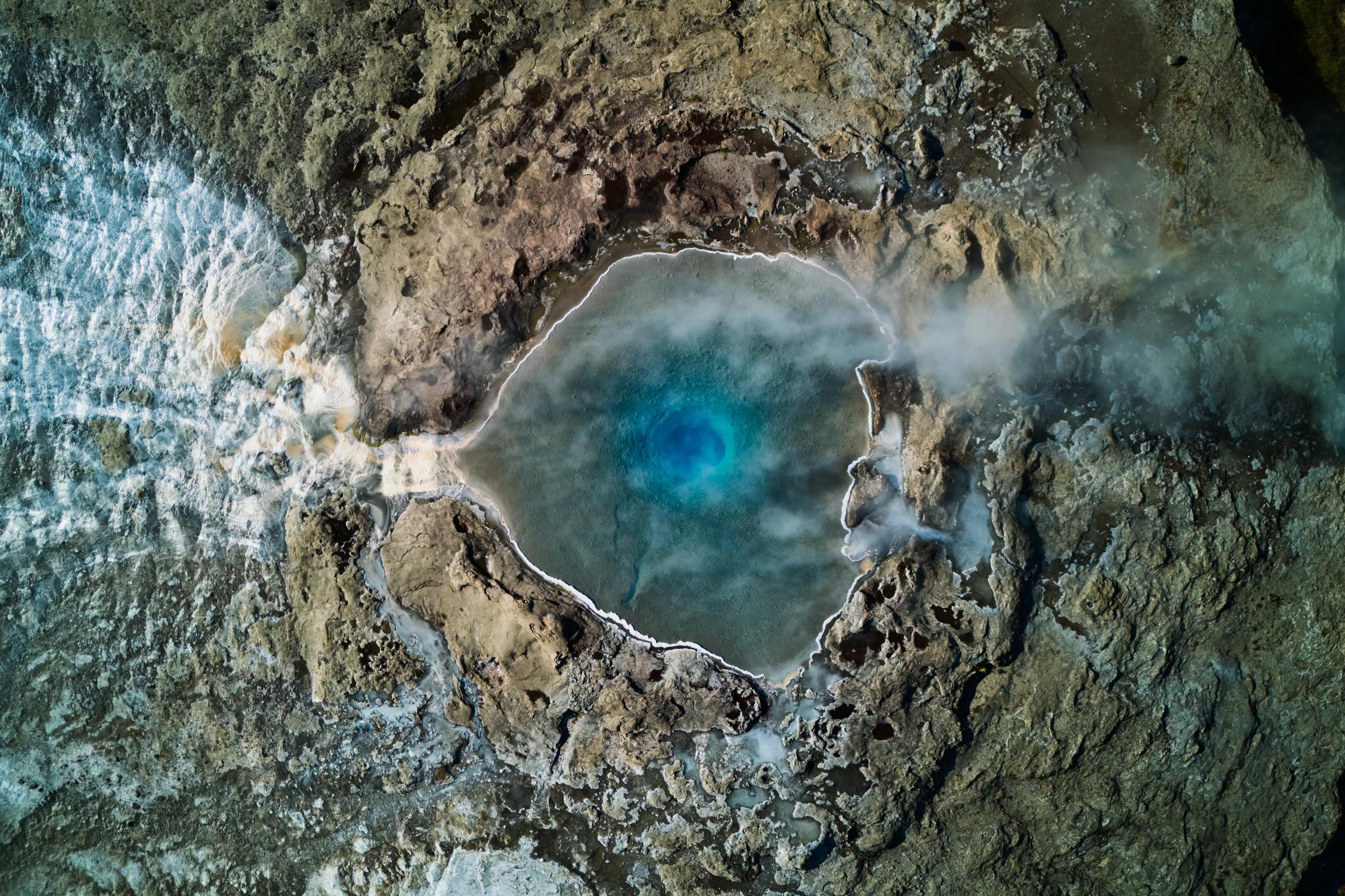 Vista aérea de una vibrante poza termal azul que humea entre un terreno rocoso, con una potente cascada a su izquierda.
