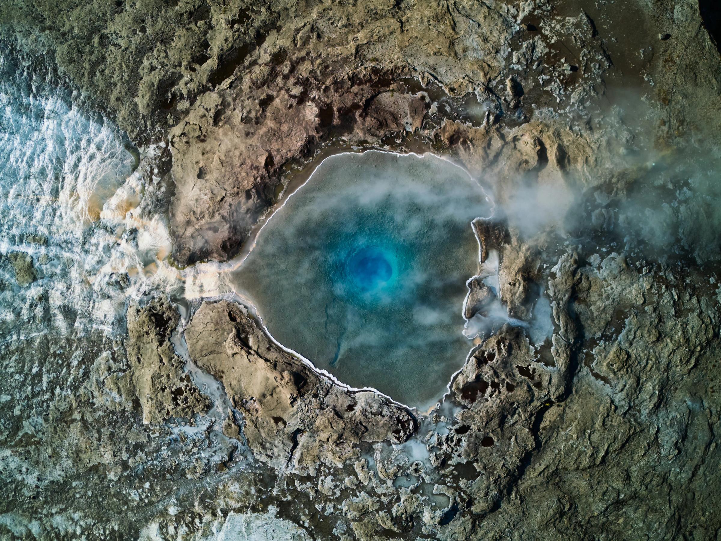 Aerial view of a bright blue, steaming geothermal pool next to a waterfall amidst rugged terrain.