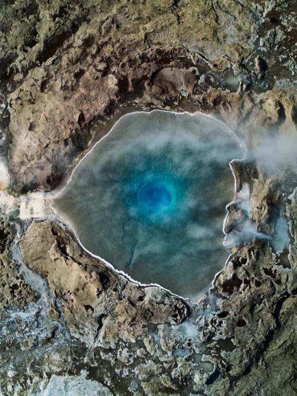 Aerial view of a vibrant blue, steaming hot spring surrounded by a rugged, rocky landscape and a waterfall.