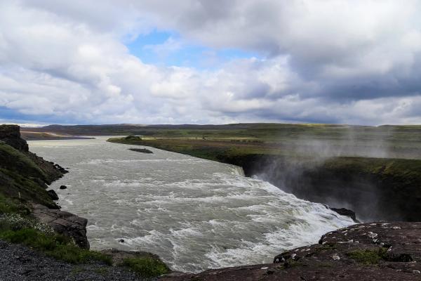 A wide river flows over a powerful waterfall, creating mist, set against flat green plains under a cloudy sky.