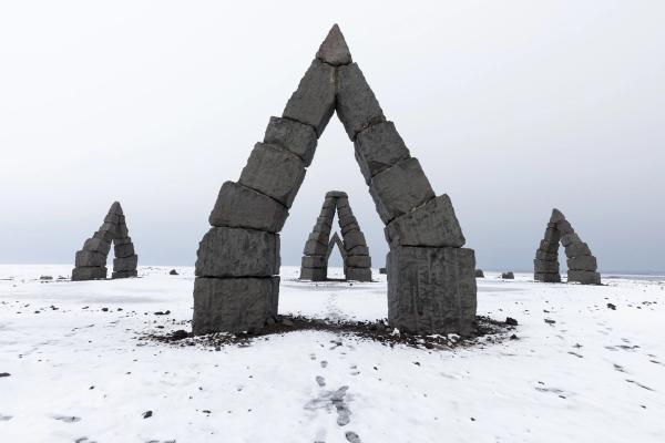 Multiple large, triangular stone arches stand in a vast, snowy landscape under a grey sky.
