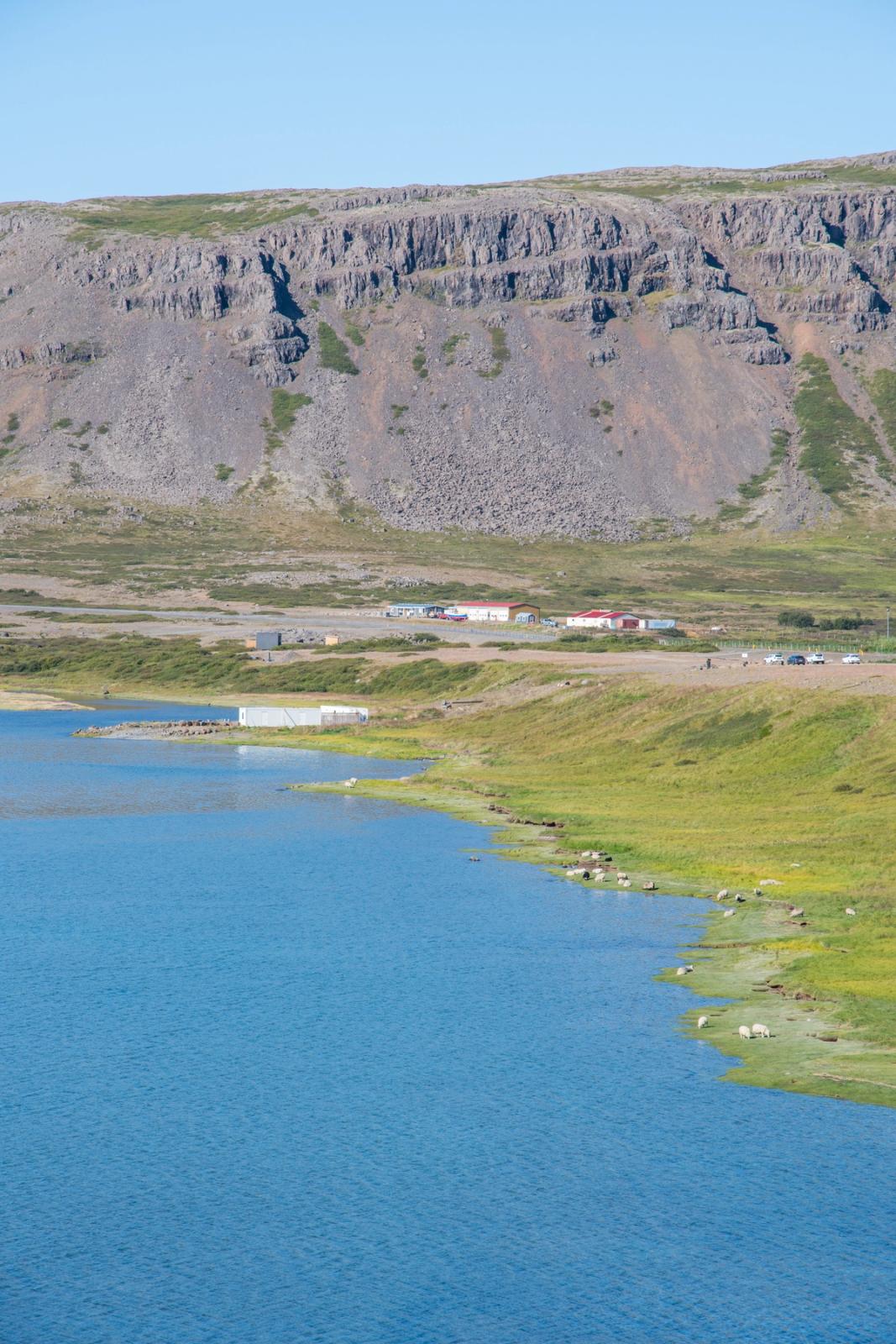 A landscape featuring a blue lake, a green shore with sheep, distant buildings, and a large rocky cliff under a clear sky.