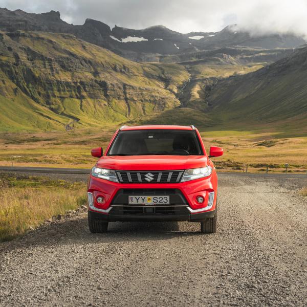 Red Suzuki Vitara SUV parked on a gravel road with green mountains and a cloudy sky in the background.