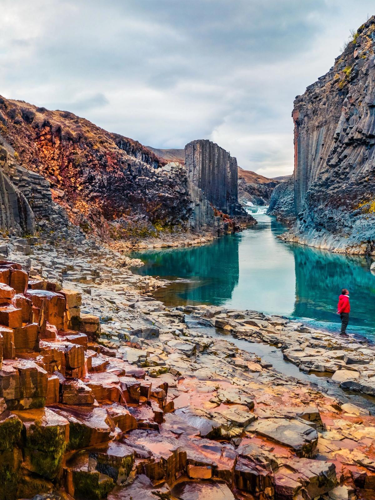 Person in a red jacket stands by a vibrant turquoise river flowing through a canyon flanked by towering columnar basalt rock formations.