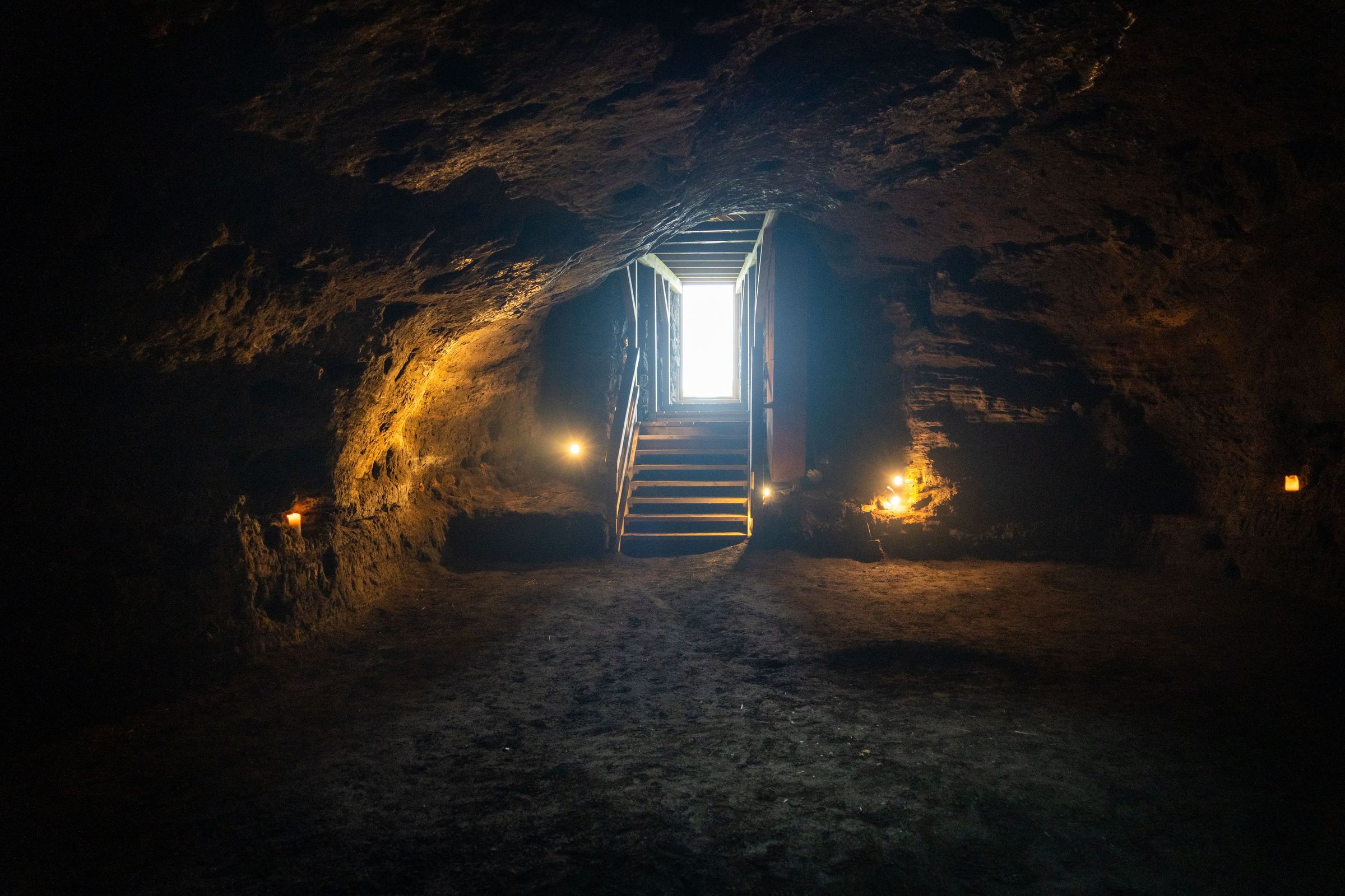 El interior de una cueva oscura con escalones que conducen a una apertura luminosa, iluminada por luces cálidas.