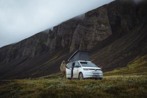 A white camper van with a pop-top tent and a person in a rugged landscape of dark mountains and waterfalls.