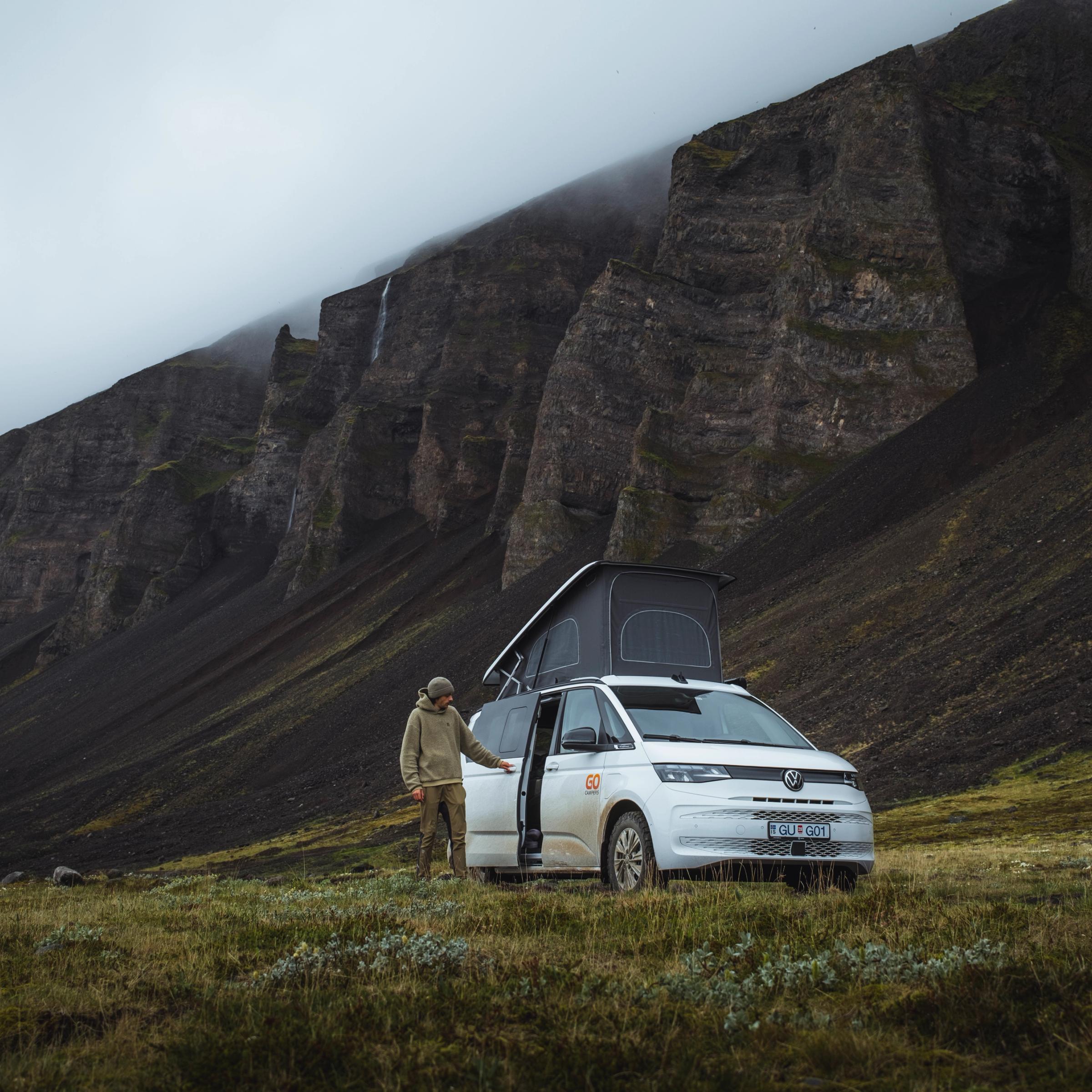 A person stands beside a white camper van with its pop-top roof extended, parked in a grassy field beneath dark, towering mountains with waterfalls under an overcast sky.