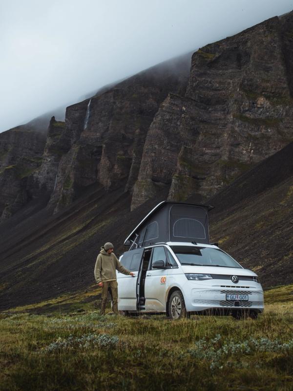 A white camper van with a pop-top tent and a person in a rugged landscape of dark mountains and waterfalls.
