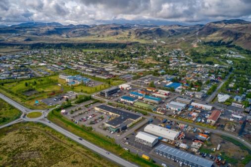 Aerial view of a town with residential and commercial buildings, roads, and a roundabout, surrounded by green fields and distant mountains under a cloudy sky.
