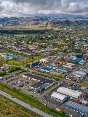 Aerial view of a town with residential and commercial buildings, roads, and a roundabout, surrounded by green fields and distant mountains under a cloudy sky.