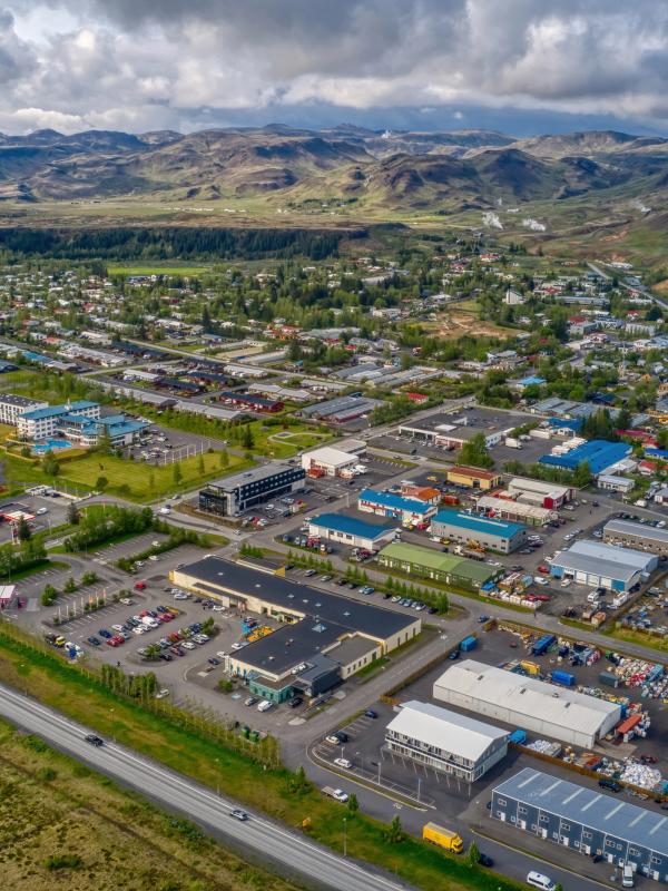 Aerial view of a town with residential and commercial buildings, roads, and a roundabout, surrounded by green fields and distant mountains under a cloudy sky.