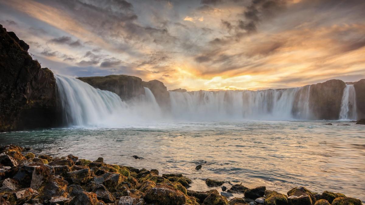 Wide waterfall at sunset with dramatic golden clouds and rocky foreground.