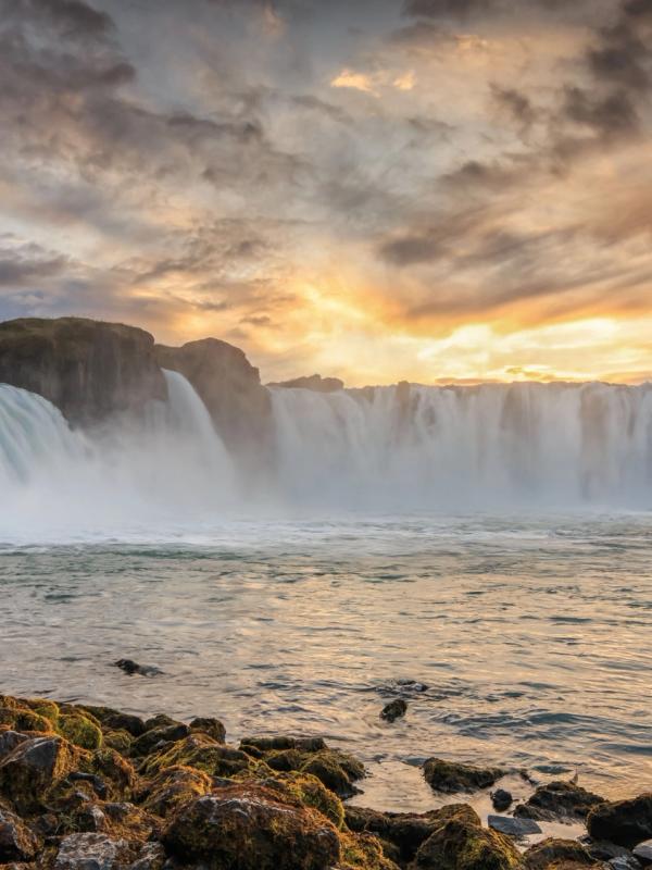 Wide waterfall at sunset with dramatic golden clouds and rocky foreground.