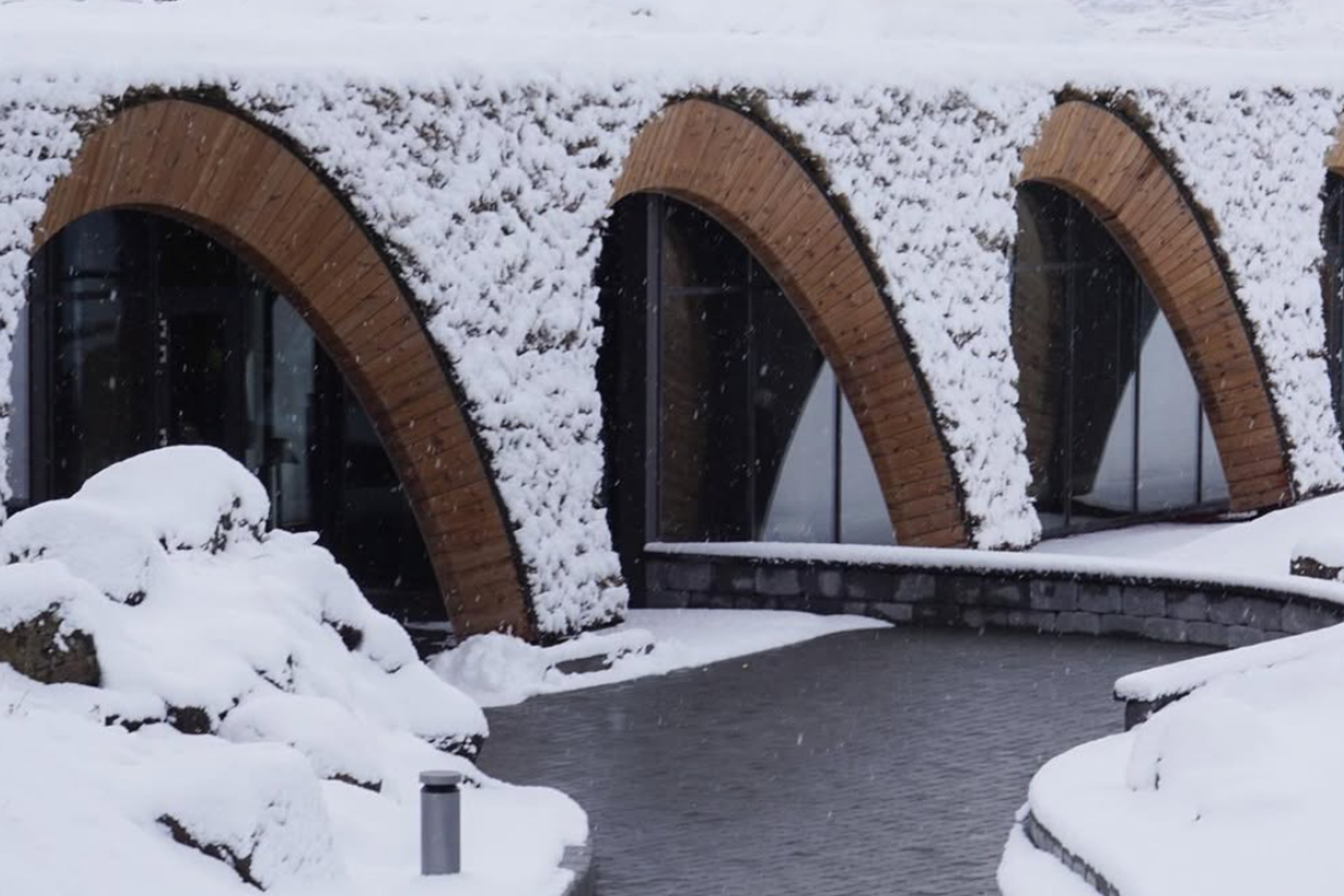 Un edificio moderno con un tejado verde cubierto de nieve y ventanas de madera arqueadas, bordeado por un sinuoso camino pavimentado en pleno invierno.