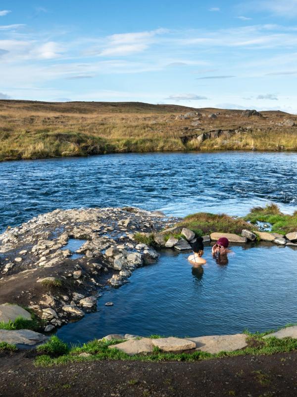 Two people bathing in a natural hot spring next to a river in a vast, grassy landscape.