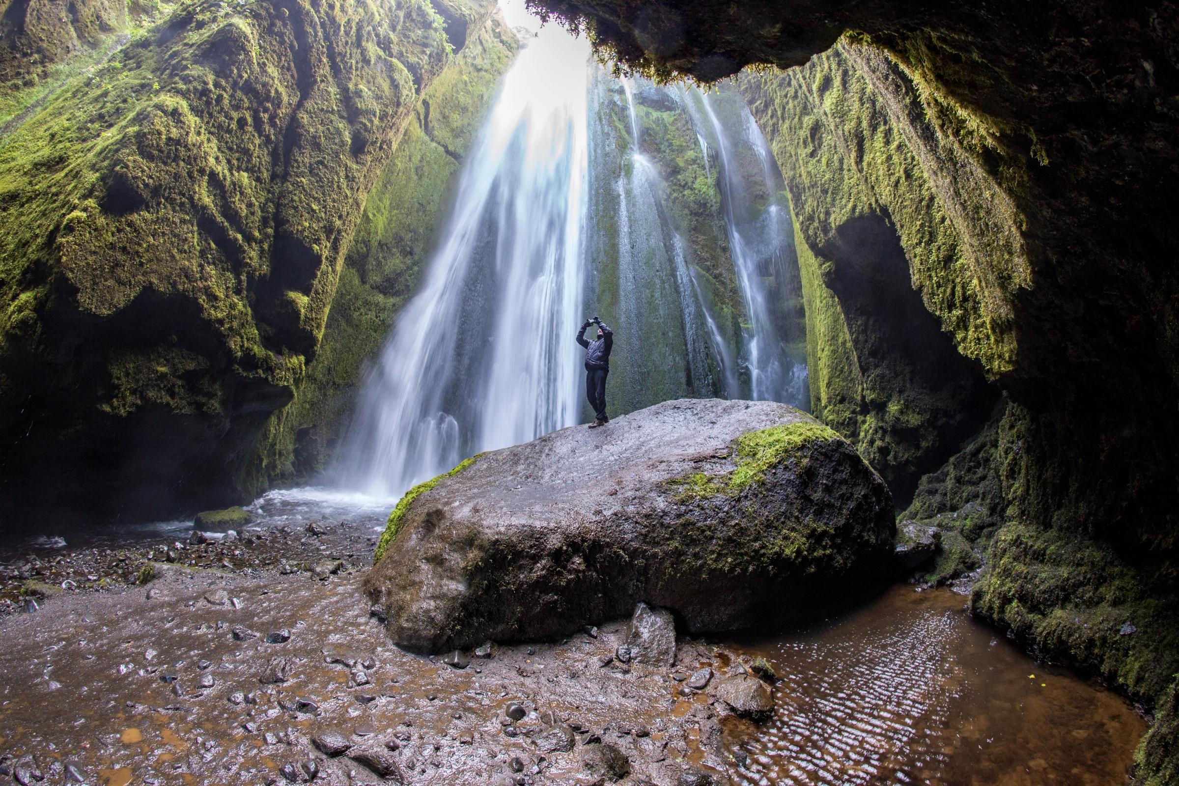 A person stands on a mossy rock inside a lush cave, looking up at a powerful waterfall streaming down from an opening in the ceiling.