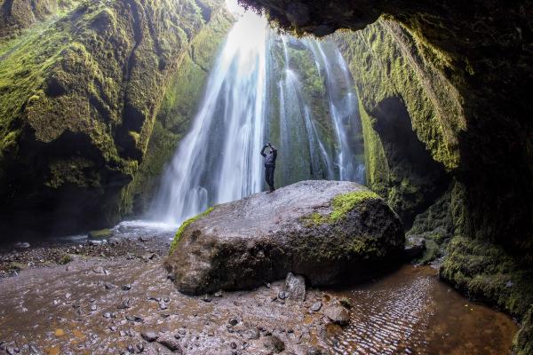 A person stands on a mossy rock inside a lush cave, looking up at a powerful waterfall streaming down from an opening in the ceiling.